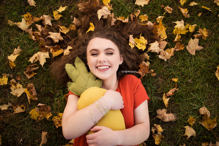 Senior girl lying down in a pile of leaves while laughing and holding a stuffed toy | Saginaw Michigan Photographer