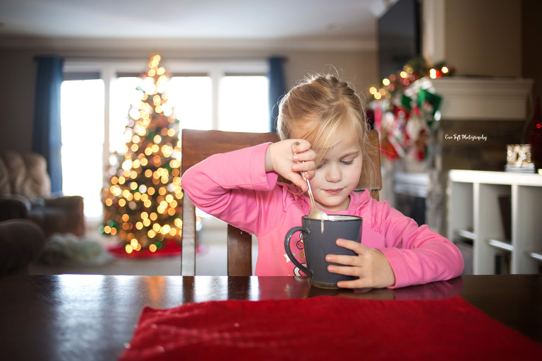 Small girl drinks hot chocolate at the table with the Christmas tree in the background | Michigan Senior Photographer