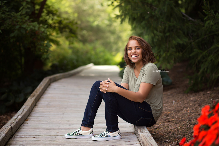 Senior girl sitting on a pathway at Dow Gardens holding her knee and smiling | Midland, Michigan Senior Photographer