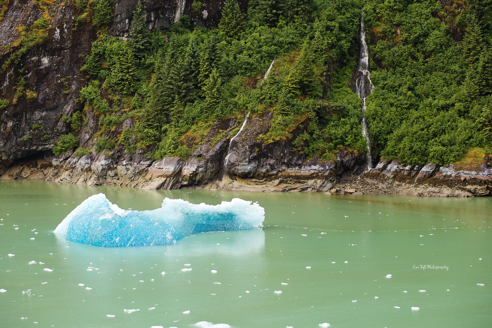 An iceberg floating down the water next to waterfalls and mountains in Alaska