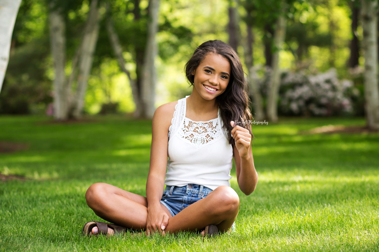 Teenage girl sitting in the grass outside holding onto her hair and smiling | Senior Photographer located in Michigan