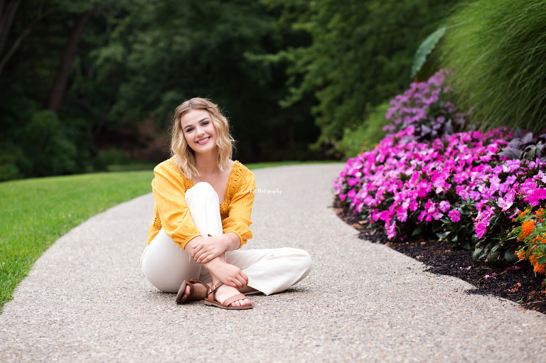 Dow High School senior sitting on a pathway at Dow Garden in Midland, Michigan next to some flowers | Senior photographer in Michigan