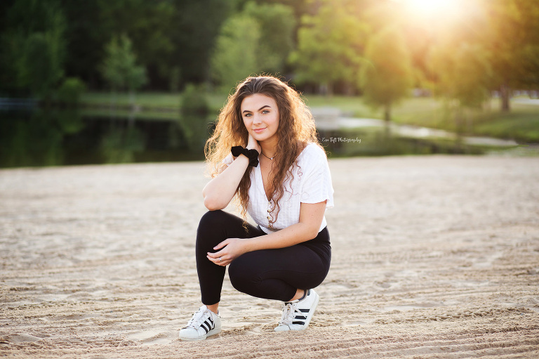 Teenage girl squatting on a beach with the sun behind her in Michigan | Senior Photographer