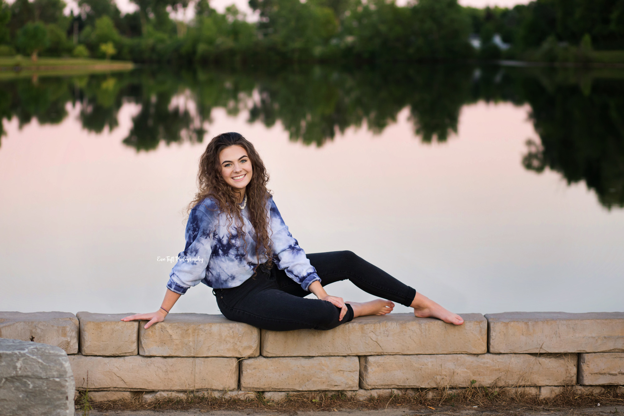 High School girl sitting on a rock wall in front of a lake in Michigan | Senior Photographer in Midland