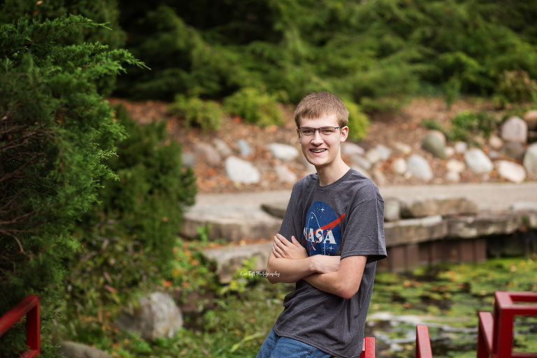 A teenage boy leaning against a railing outside by a small pond at Dow Gardens | Senior Photographer in Midland, Michigan