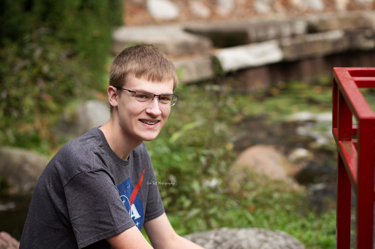 A senior guy smiles at the camera while sitting on rocks by a stream | Seniot Photographer in Midland, Michigan