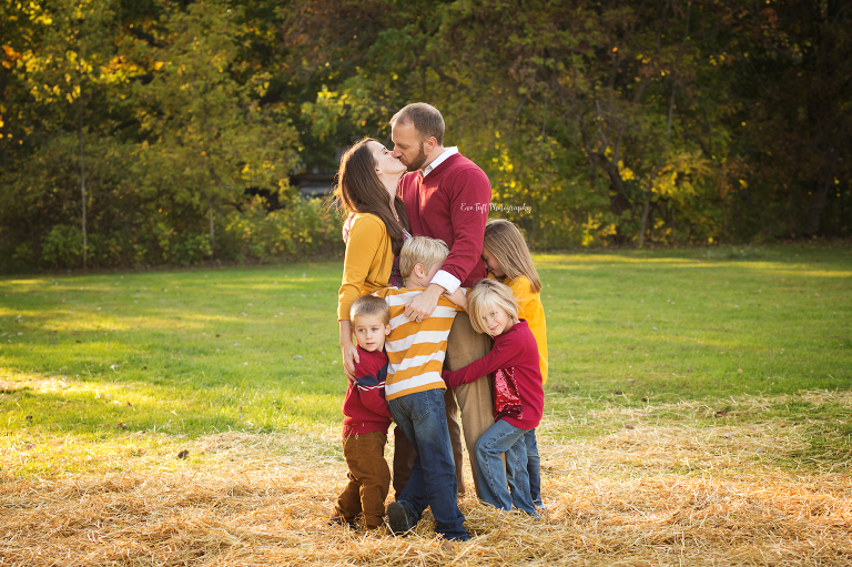 A couple kiss while standing and their four young kids hug them | Michigan Photographer