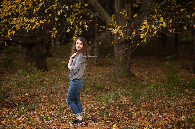A high school girl poses outside in the fall leaves under a tree at the Chippewa Nature Center | Senior Photographer in Midland, Michigan