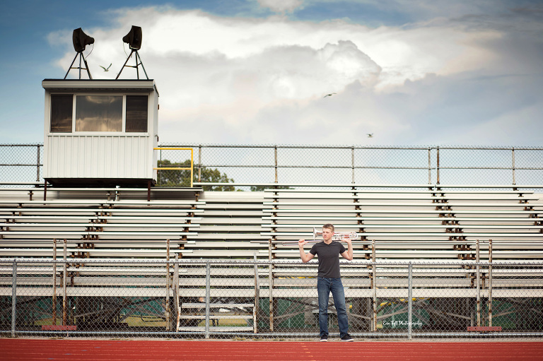 A teenage guy holds his trombone across his shoulders in front of football stands | Senior Photographers in Midland, Michigan