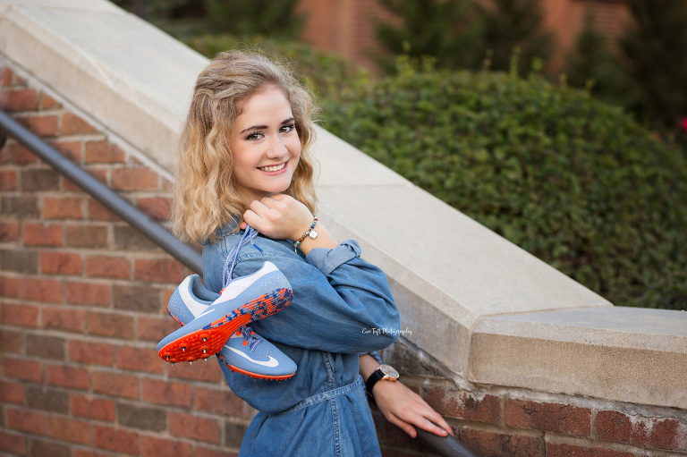 A girl holds her pair of running shoes in her hands as her props as she looks over her shoulder | Michigan Senior Photographer