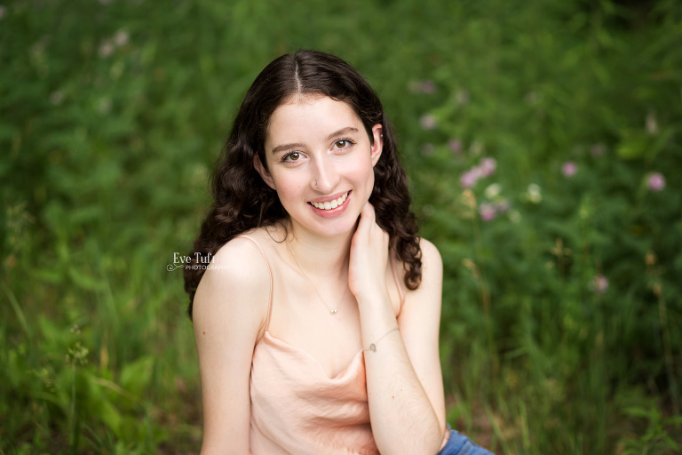 A teenage girl sits in wildgrass while smiling at the camera with her hand on her neck in City Forest | Midland, Michigan Senior Photographers