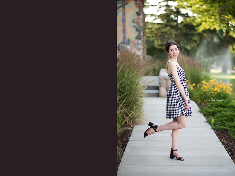 A senior girl faces the camera at the courthouse on the sidewalk with one leg lifted up in a cute pose | Michigan senior photographer in Midland