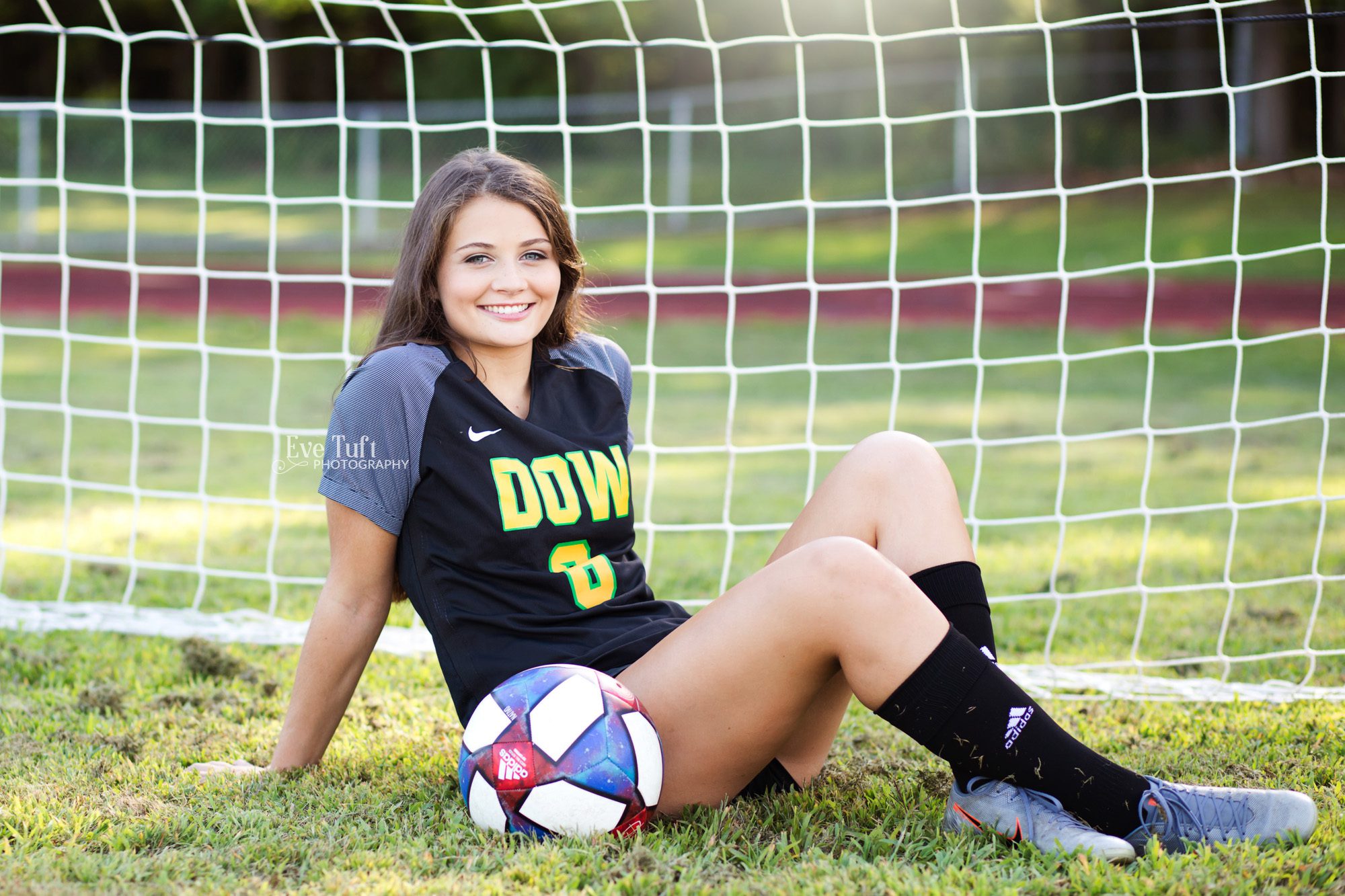  A sports picture of a teenage girl sitting in front of a goal with a soccer ball on the ground next to her | Senior Photographer in Midland, Michigan