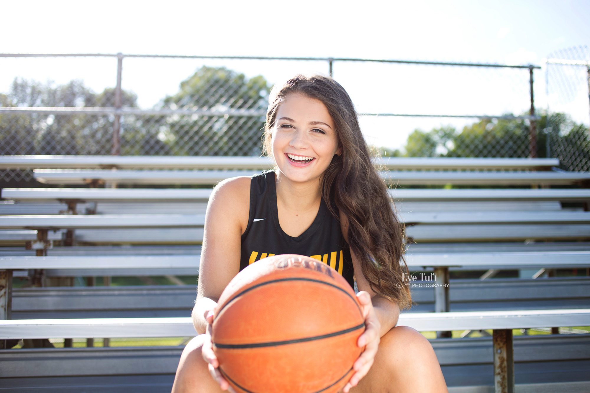 A senior girl holds a basketball on the bleachers at H. H. Dow High School in Midland, MI | Senior Photographers in Midland, Michigan