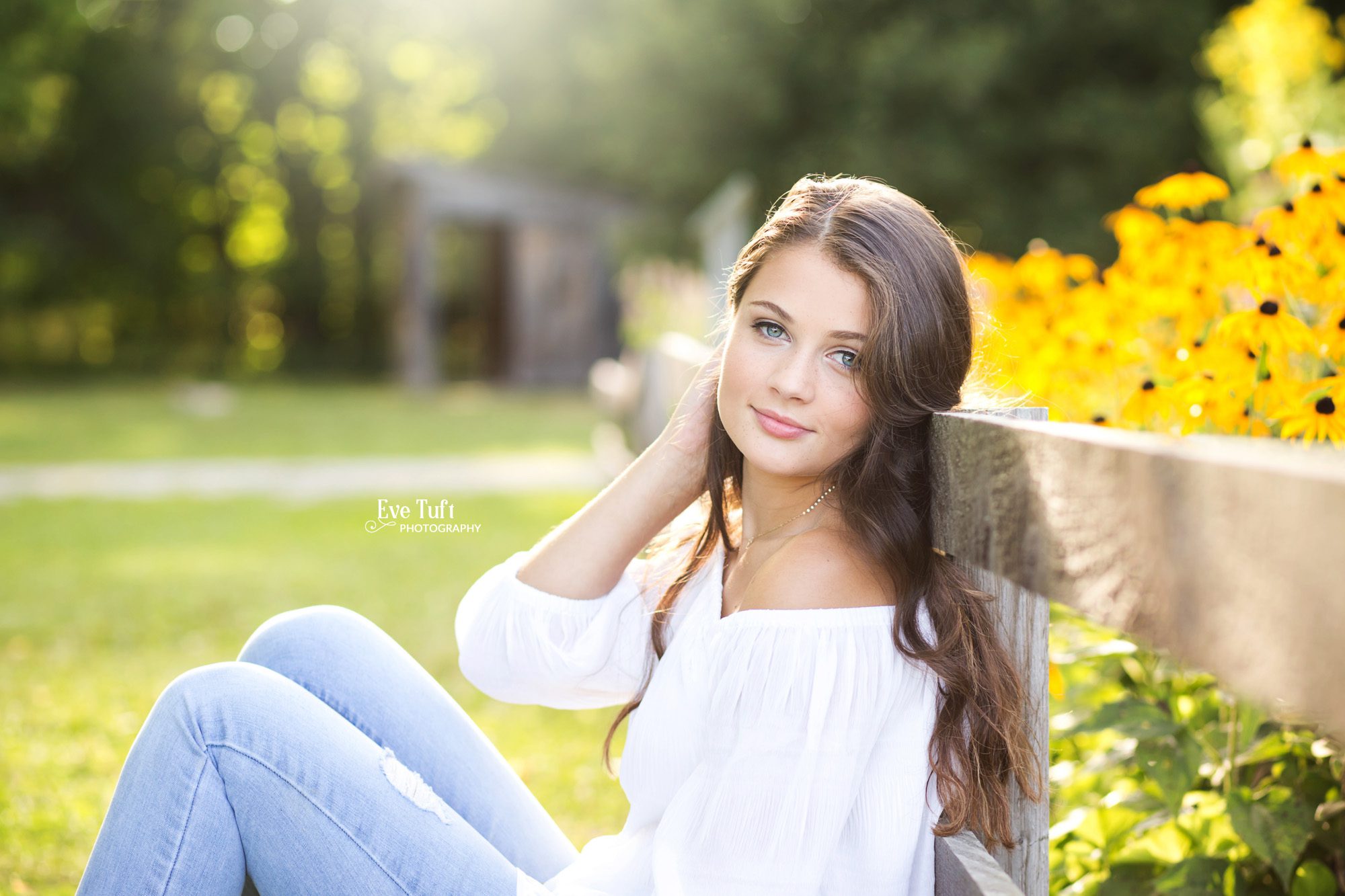 A senior girl leans up against a wooden fence while sitting on the ground at the Chippewa Nature Center in Midland, MI