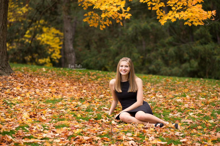 Beautiful senior girl sits in a pile of autumn leaves outside on a hill for her senior pictures | senior Photographers in Midland, Michigan