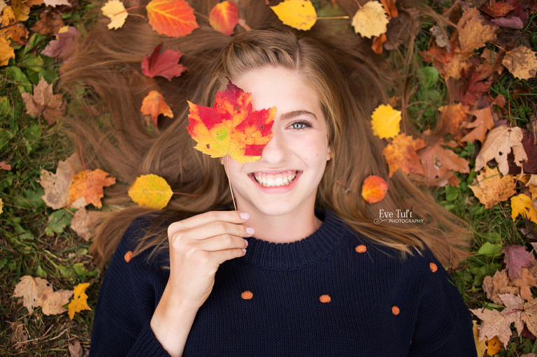 A teenage girl lies on the ground while holding up a colorful fall leaf to her eye | Senior Photographers in Midland, Michigan